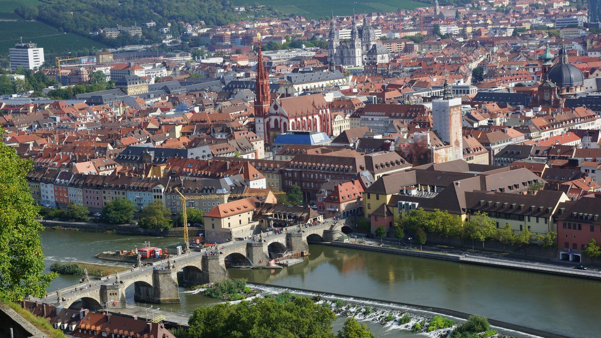 Blick auf eine Stadt im Sommer. Im Vordergrund ein breiter Fluss, über den eine alte Steinbrücke führt.