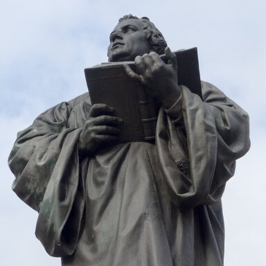 Eine Statue Martin Luthers, der ein offenes Buch hält. Im Hintergrund ein blauer Himmel mit Schleierwolken.