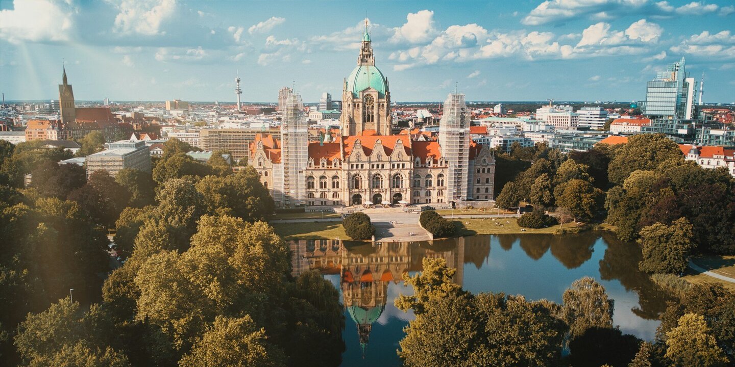 Blick auf das hannoversche Rathaus und die dahinter liegende Stadt. Im Vordergrund Bäume und ein Teich.